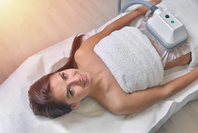 Woman receiving CoolSculpting treatment on abdomen while lying on a medical bed, using a non-invasive fat reduction device.