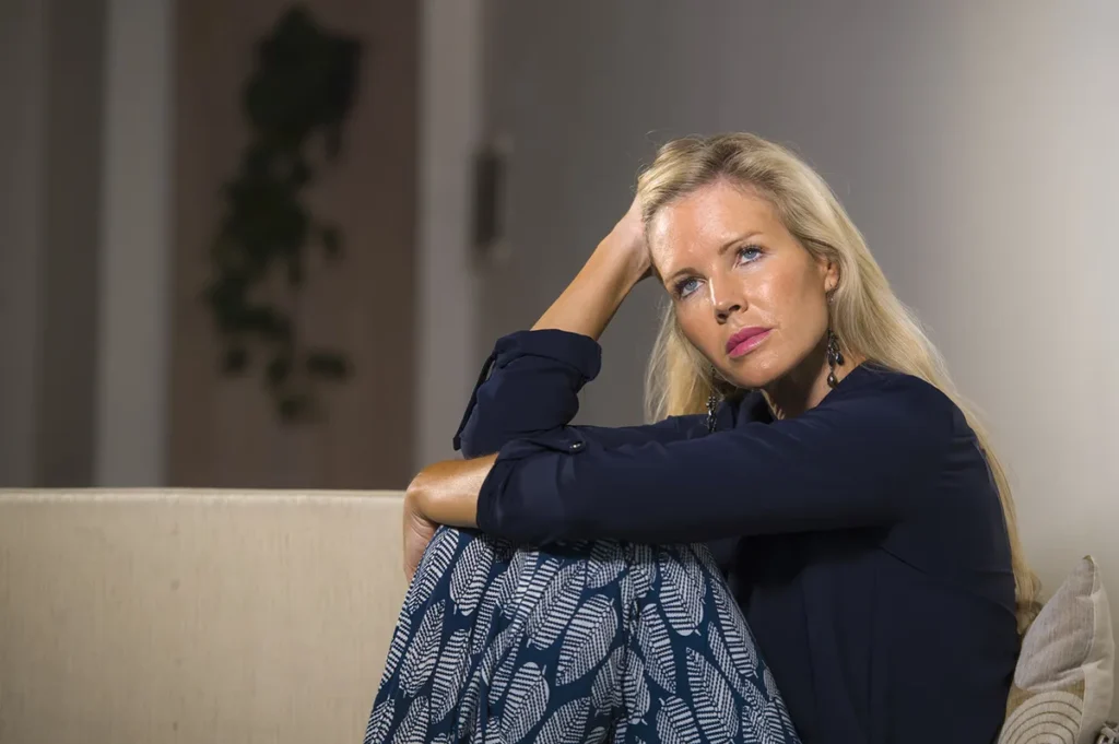 A fair-skinned woman with long blonde hair and blue eyes sits on a couch, looking pensive or sad, with one hand resting on her head. She is wearing a dark long-sleeved top and patterned pants.