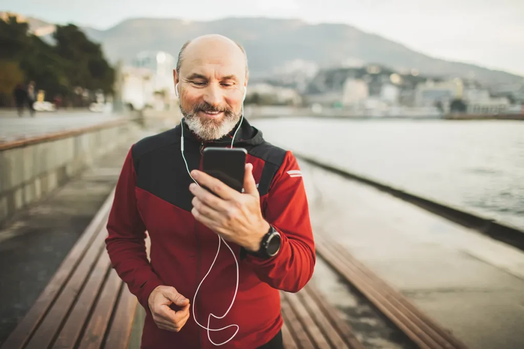 A smiling, balding man with a beard and earbuds looks at his phone while walking outdoors next to a body of water with mountains in the background. He is wearing a red, black, and gray athletic jacket and a smartwatch.