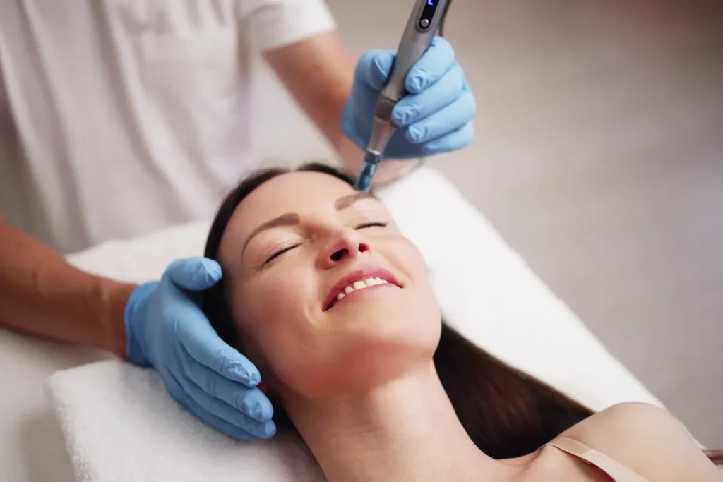 A woman with long dark hair smiles while receiving a microneedling facial treatment on her forehead, performed by a professional wearing blue gloves and using a handheld device.