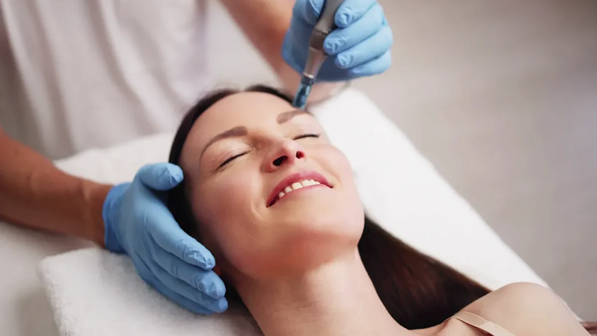 A woman with long dark hair smiles while receiving a microneedling facial treatment on her forehead, performed by a professional wearing blue gloves and using a handheld device.