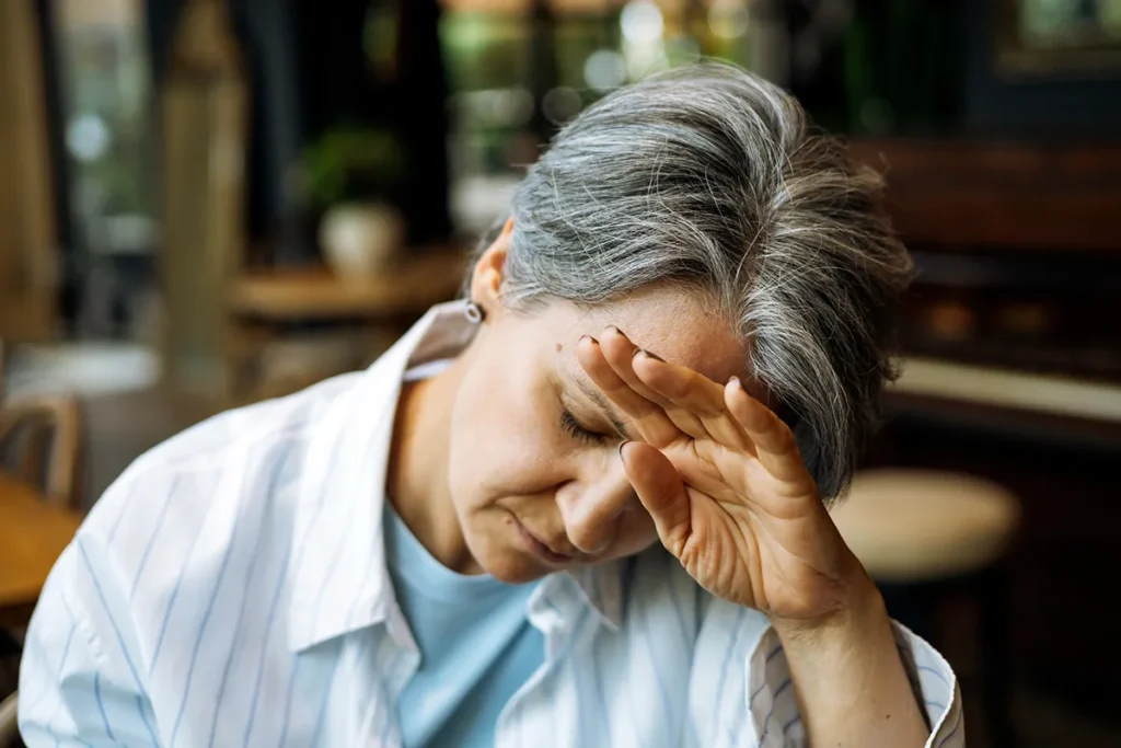 A woman with short, graying hair holds her hand to her forehead, looking down with a pained or tired expression. She is wearing a light blue shirt under a striped white collared shirt.